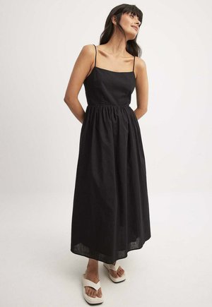 Abito maxi nero con spalline sottili, busto aderente e vita arricciata. Tessuto liscio con gonna fluente, abbinato a ciabatte bianche.