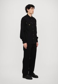 Camisa negra de botonadura con dos bolsillos en el pecho, detalles en dorado y mangas largas, combinada con pantalones y zapatos negros.
