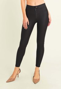 Leggings negros ajustados con cintura alta, cierre frontal con cremallera y costuras verticales en contraste. La tela tiene una textura suave y elástica.