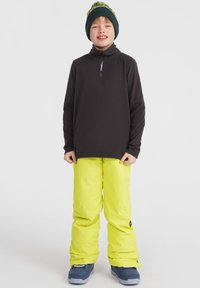 Haut noir à manches longues avec half zip, pantalon de neige jaune vif et bottes de neige bleues. Complété par un bonnet vert avec un pompon.