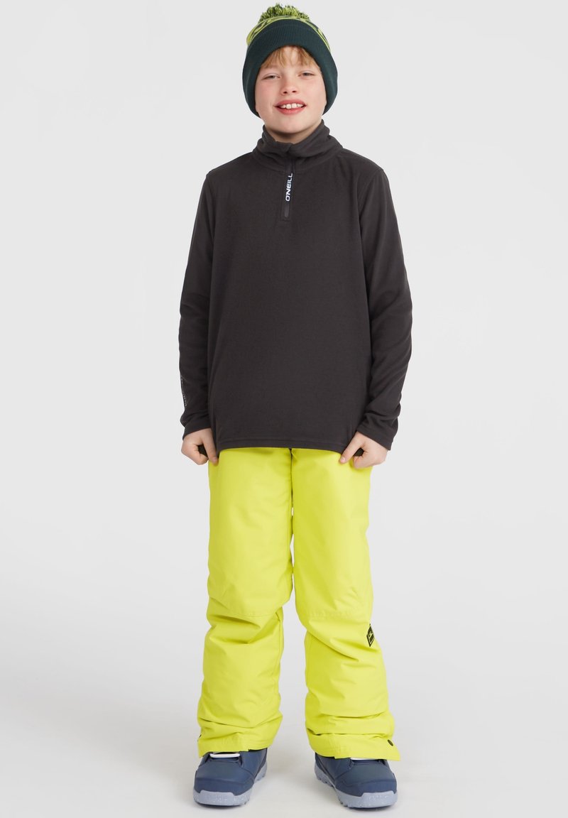 Haut noir à manches longues avec half zip, pantalon de neige jaune vif et bottes de neige bleues. Complété par un bonnet vert avec un pompon.