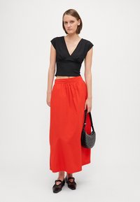 Fekete, V-nyakú, cropped top, amelyet egy lebegő piros maxi szoknyával párosítanak. A modell egy kis fekete táskát tart ezüst szegecsekkel, fekete cipőt viselve.