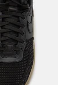 Närbild av svart sneakers med texturerad tåhuvud, svarta snören och beige sula på vit bakgrund.