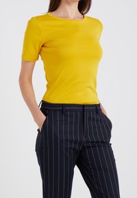 Žluté tričko s krátkým rukávem a kulatým výstřihem, vyrobené z hladkého materiálu, spárované s tmavomodrými kalhotami s pruhy a bočními kapsami.