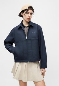 Νεαρός με μπεζ καπέλο φορεμένο ανάποδα, μπλε ναυτικό ζακέτα Carhartt με φερμουάρ, ριγέ πόλο πουκάμισο και μπεζ πλισέ φούστα, ποζάρει μπροστά σε απλό φόντο.