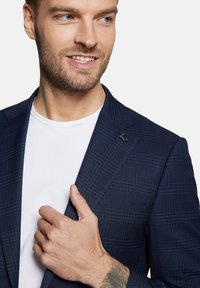 Granatowa marynarka w kratę z teksturowanej tkaniny, z ostrymi klapami i subtelną przypinką z logo. Nosi na sobie białą koszulę, ręka spoczywa na klapie.