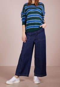 Gestreepte groene en blauwe gebreide trui gecombineerd met donkerblauwe wijde jeansbroek en witte sneakers. Zachte textuur, ontspannen pasvorm, met opvallende zakken.