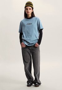 Lichtblauw T-shirt met grafische print, gedragen over een zwart longsleeve shirt. Losse grijze jeans. Zwarte schoenen en een meerkleurige beanie.