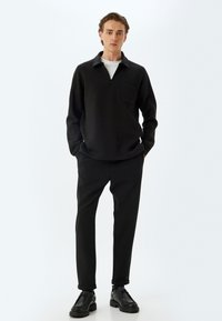 Pullover nero a zip a quarti con una tasca singola sul petto, abbinato a pantaloni neri affusolati e scarpe nere con lacci, su uno sfondo bianco.