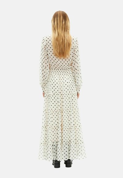 Lang weißes, durchsichtiges Kleid mit schwarzem Herzmuster. Es verfügt über gestufte Rüschen, lange Ärmel und eine taillierte Passform. Kombiniert mit schwarzen Stiefeln.