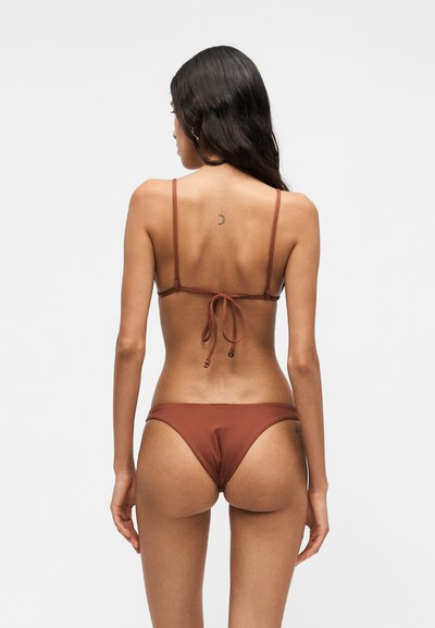Mujer con cabello largo y oscuro lleva un bikini color óxido, de espaldas, mostrando la espalda y un pequeño tatuaje de luna creciente en la parte superior de la espalda.