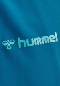 Turquoise stof met het "hummel" logo in lichtblauw. Het materiaal heeft een gladde textuur, met een gestileerd bij-embleem naast de tekst.