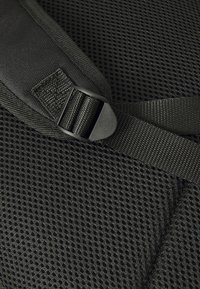 Black adjustable nylon strap with plastic buckle over breathable mesh fabric, likely part of a backpack or carrier.