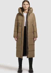 Dlhý hnedý puffový kabát s kapucňou, so zipsom na prednej strane, bočnými vreckami a prešívanou textúrou, skombinovaný s čiernymi vysokými čižmami po kolená.