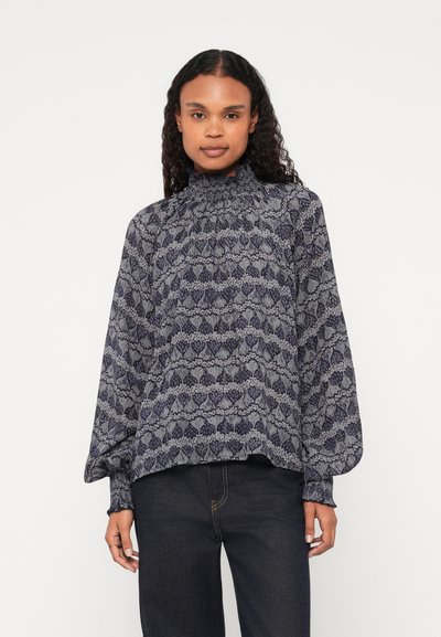 Langsleeve blouse met een hoge gerimpelde hals, voorzien van een marineblauwe en lichtgrijze bladenpatroon op een lichte stof. Gecombineerd met donkere jeans.