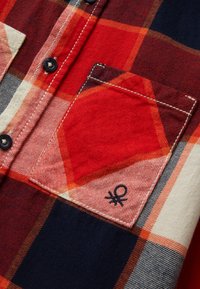 Camicetta a quadri rossa, arancione, blu navy e crema con tasca, bottoni neri e piccolo logo ricamato nero vicino all'angolo inferiore.