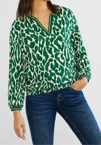 Blusa a stampa leopardata verde e crema con scollo a V, polsini elasticizzati e vestibilità rilassata. Indossata con jeans blu, che mostra un look informale.