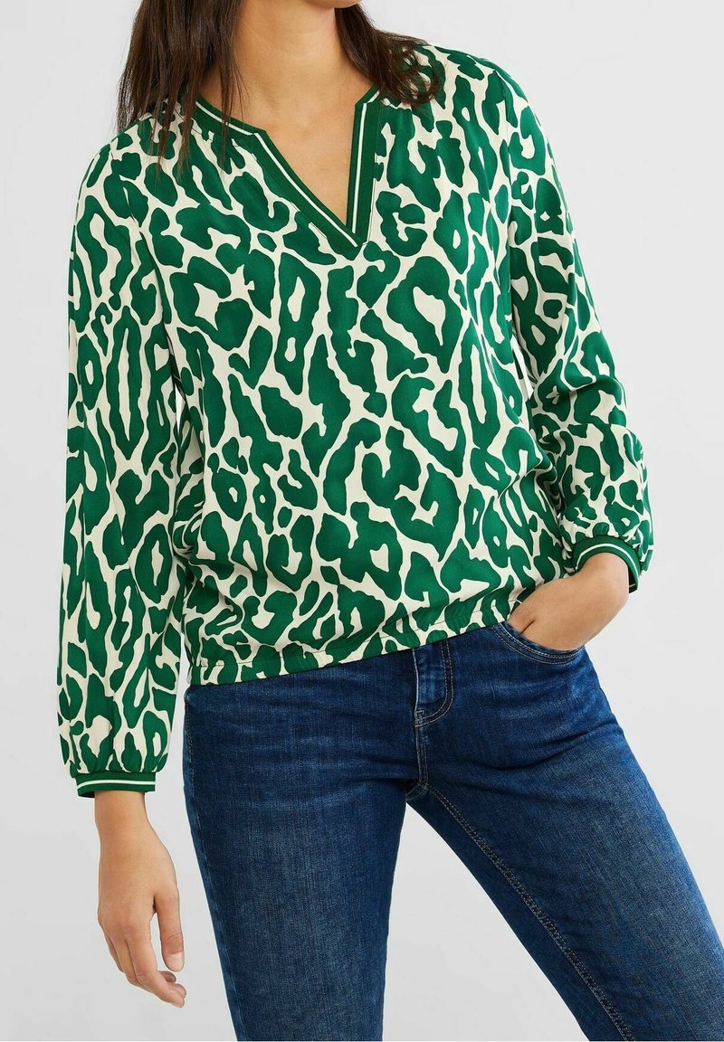 Blusa a stampa leopardata verde e crema con scollo a V, polsini elasticizzati e vestibilità rilassata. Indossata con jeans blu, che mostra un look informale.