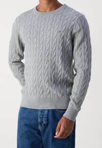 Grauer Kabelstrickpullover mit Rundhalsausschnitt, gerippten Bündchen und Saum. Kleines Logo-Detail in der Nähe der Brust. Getragen mit blauen Jeans.