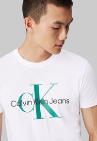 White cotton t-shirt featuring large teal and black "Calvin Klein Jeans" graphic print, round neckline, and short sleeves.