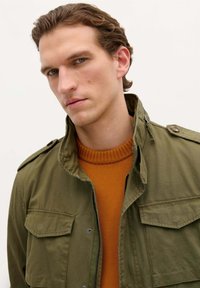 Olijf groene militaire stijl jas met drukknoopzakken over een gestructureerde oranje gebreide trui, met een ritsluiting en verhoogde kraag.