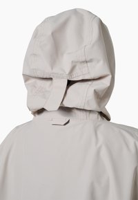 Jasna beżowa wodoodporna kurtka widziana od tyłu, z naszytym kapturem, detalami przeszyć i regulowanym paskiem.