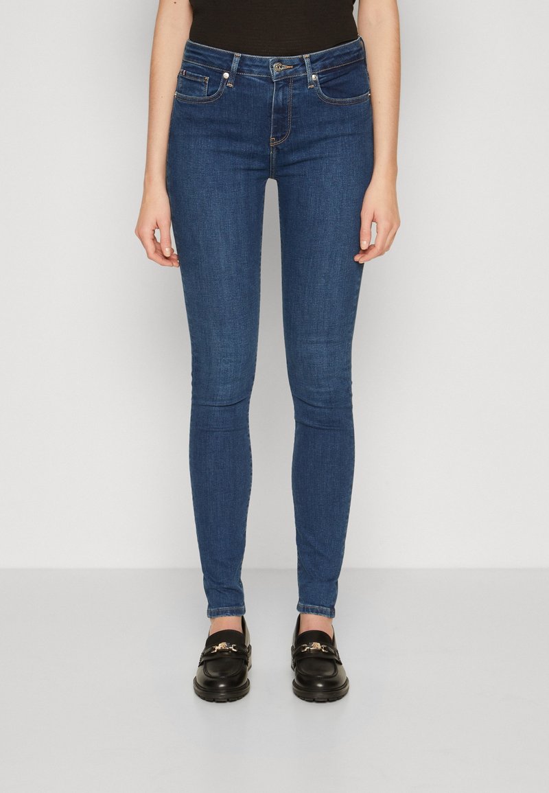 Tmavě modré úzké džíny s hladkou denimovou texturou, obsahující pět kapes a zapínání na přední knoflík. Měly na sobě černé mokasíny.