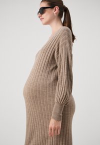Beige bordás kötött kismama ruha hosszú, szoros ujjal és V-alakú nyakkivágással, textúrált mintázattal. A sziluett testhez simuló.