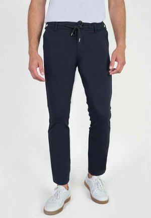 Marineblauwe slim-fit broek met een trekkoord in de taille, voorzien van zijzakken en strakke lijnen. Draagt met witte sneakers en een casual top.