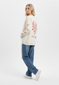 Felpa color crema con grafiche di cuori rossi sul retro, schizzi neri sulle maniche, abbinata a pantaloni blu testurizzati e sneaker bianche.