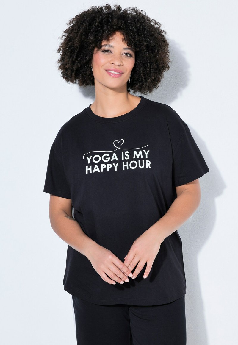 Femme aux cheveux bouclés portant un t-shirt noir avec le texte blanc "YOGA IS MY HAPPY HOUR", debout devant un fond clair, souriante.