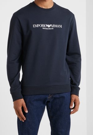Man draagt een marineblauwe Emporio Armani sweatshirt met witte logo-tekst en donkerblauwe spijkerbroek, staand tegen een effen achtergrond.