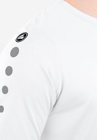 White long-sleeve shirt with a smooth texture, featuring a black logo on the shoulder and black dotted accents along the sleeve.