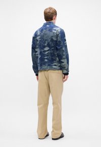 Chaqueta de mezclilla en tonos azules con un patrón texturizado, que presenta un cuello y puños con botones, combinada con pantalones beige.