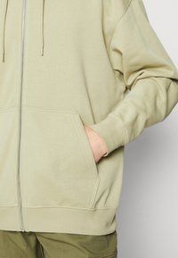 Sudadera con cremallera de color verde claro, hecha de material suave, con un bolsillo tipo canguro, puños acanalados y una textura suave. Diseño sencillo.