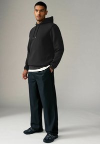 Zwarte hoodie met textuur en een voorzak, gecombineerd met losse zwarte broeken en zwarte sportschoenen. Een wit T-shirt is zichtbaar eronder.