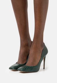 Jambes à la peau foncée portant des talons aiguilles pointus en cuir serpent de couleur vert foncé sur fond blanc.