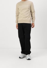 Sudadera beige con texto de logo blanco, combinada con pantalones negros de pierna ancha y zapatillas deportivas gris claro, de pie sobre un fondo liso.