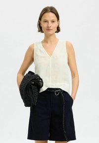 Suknja bez rukava od lana u boji off-white s V izrezom. Uparena s tamnoplave kratke hlače s užetom. Drži crnu heklanu torbicu.