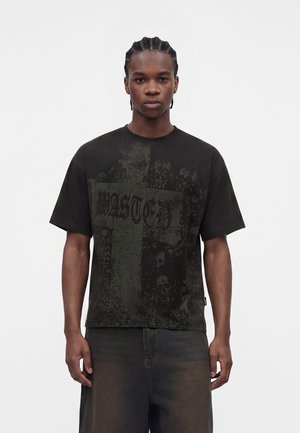 Junger Mann mit geflochtenem Haar, der ein schwarzes oversized T-Shirt mit einem gotischen Kreuz und dem Schriftzug „WASTED“ trägt, kombiniert mit lockeren, dunklen Hosen.