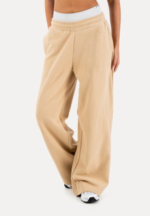 Pantalones deportivos beige de pierna ancha con cintura elástica blanca y zapatillas, usados por una persona recortada a la altura de la cintura, de pie contra un fondo blanco.
