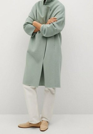Personne portant un manteau oversize vert clair, un pantalon blanc et des mocassins beiges, debout les bras croisés devant un fond uni.