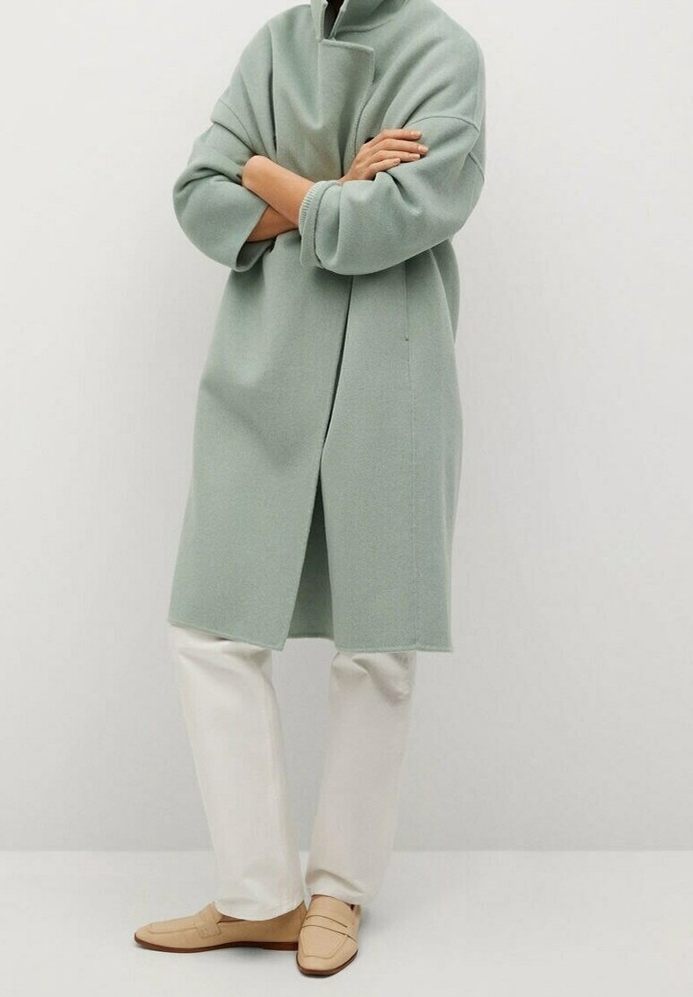 Personne portant un manteau oversize vert clair, un pantalon blanc et des mocassins beiges, debout les bras croisés devant un fond uni.