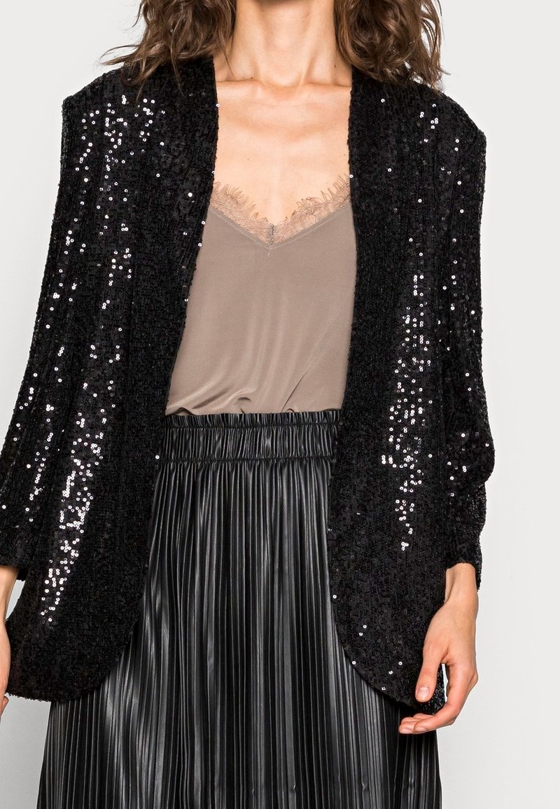 Femme portant un blazer noir à sequins sur un caraco taupe bordé de dentelle et une jupe noire plissée taille haute.