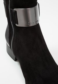Bottines en suède noir avec un accent métallique portant la marque "Calvin Klein", fermeture éclair sur le côté et un talon épais pour la stabilité.