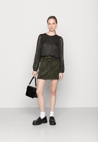 Blusa preta transparente com riscas verticais douradas, saia de cortiça verde-oliva com botões à frente, sapatos de plataforma pretos e uma mala preta.