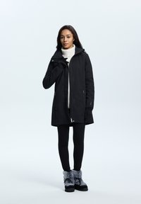 Veste noire imperméable avec un col haut, fermeture éclair à l'avant et poches latérales, associée à un pull à col roulé blanc et à des leggings noirs, ainsi qu'à des bottes en fourrure grises.