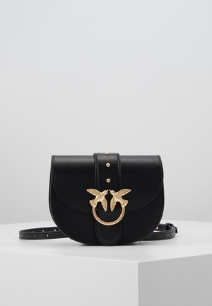 Sac bandoulière en cuir noir avec un fermoir doré en forme d’oiseau et des clous dorés, présenté sur un piédestal blanc sur fond gris.