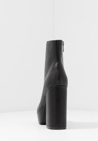 Botín de piel negra hasta el tobillo con un tacón alto y grueso, y un detalle de cremallera en la parte trasera, que presenta una textura suave y un diseño aerodinámico.