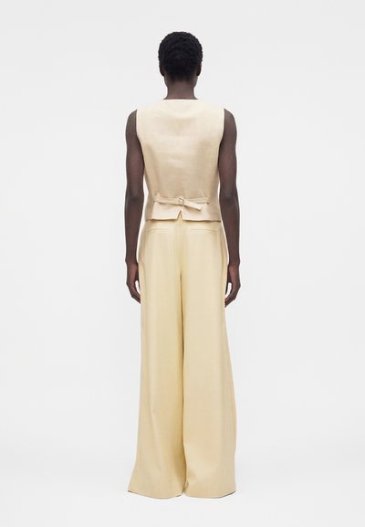 Άτομο με κοντά σγουρά μαλλιά που φοράει μπεζ αμάνικο γιλέκο με αγκράφα στην πλάτη και εκρού παντελόνια φαρδιάς γραμμής, στέκεται γυρισμένο ανάποδα.